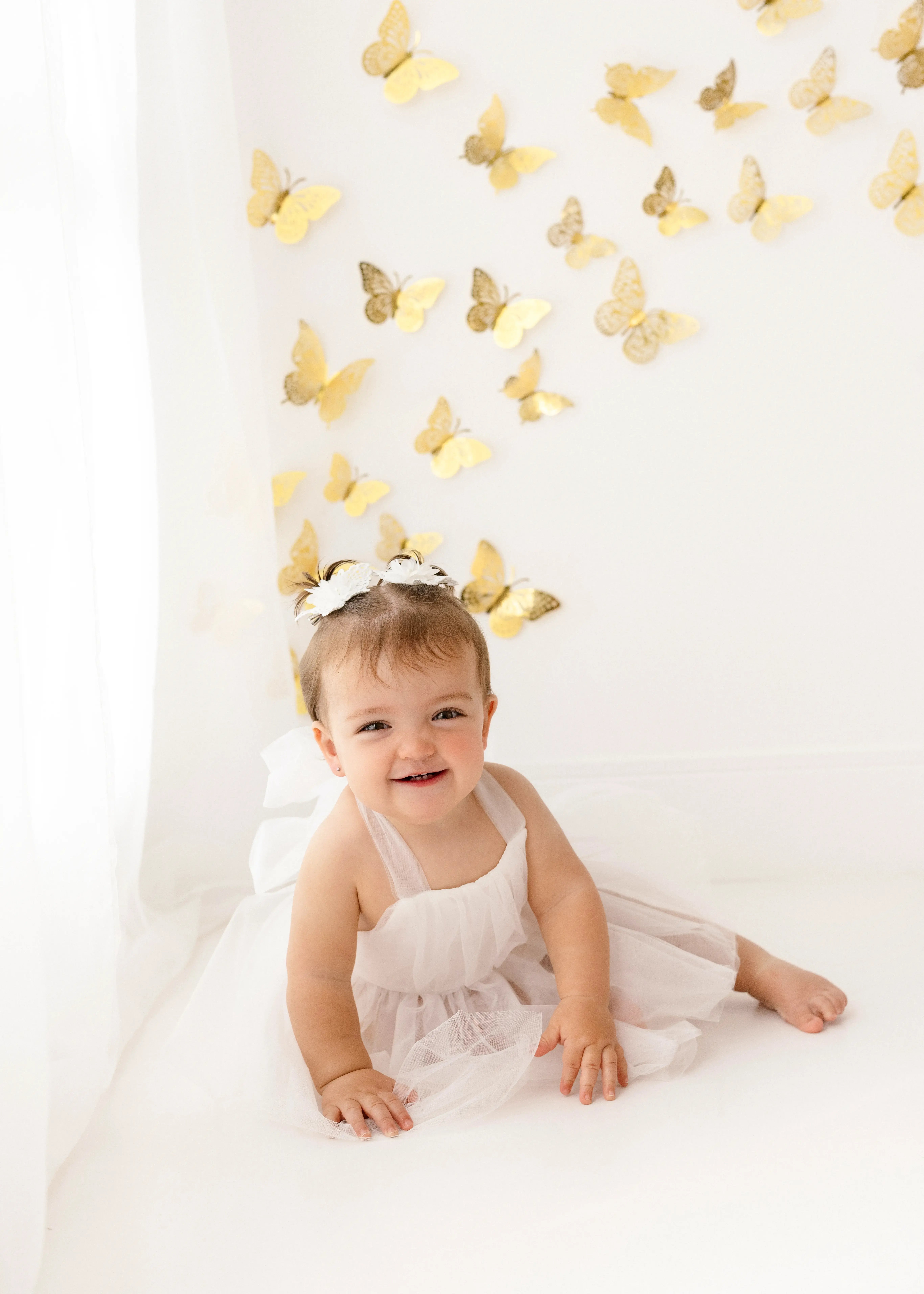 Adorable toddler girl in a white tulle dress with floral hair accessories, sitting on a white floor beneath a wall decorated with golden butterflies.